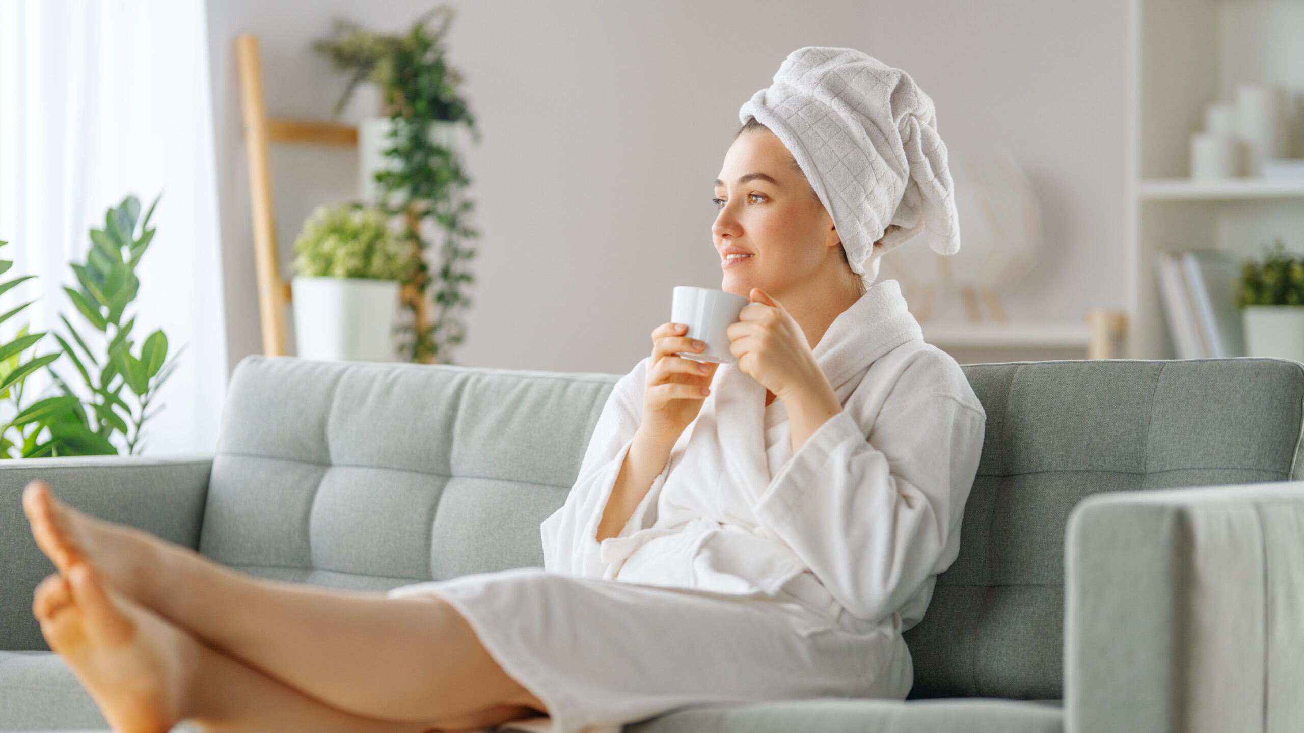 A imagem mostra uma pessoa sentada em um sofá com os pés para cima, vestindo um roupão branco e uma toalha enrolada na cabeça. A pessoa está segurando uma caneca e parece estar em uma postura relaxada. O rosto da pessoa está obscurecido por um retângulo marrom, provavelmente por razões de privacidade. Há plantas e decoração de casa ao fundo.