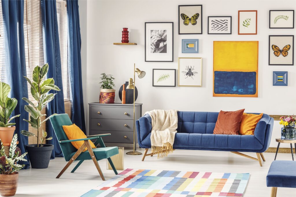 Imagem retratando um ambiente com decoração colorida. A decoração é composta por um tapete com quadriculado colorido, sofá azul, cadeira de descanso verde, vasos de planta e diversos quadros com temas de folhas e borboletas