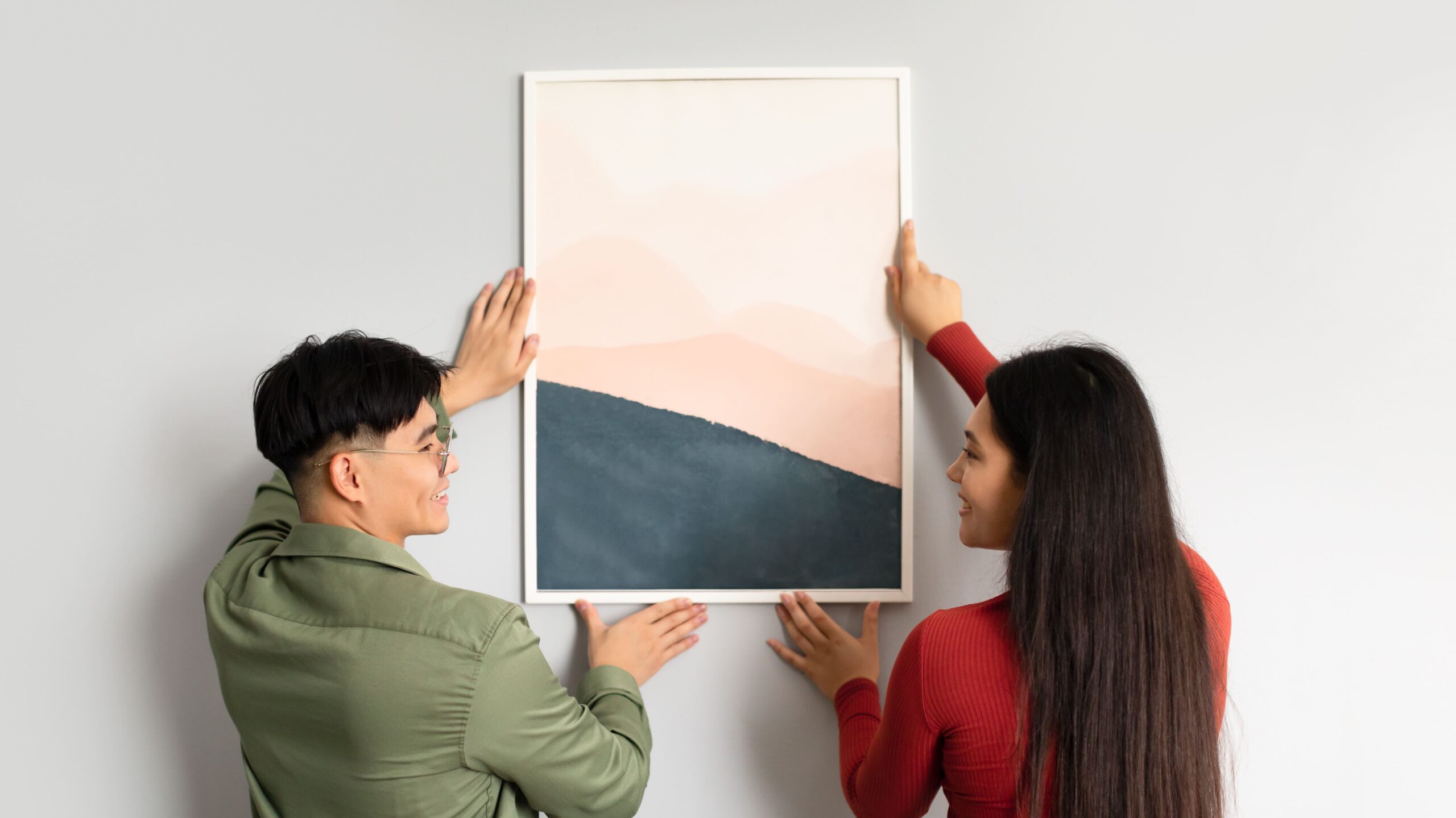Homem e mulher, jovens, pendurando um quadro decorativo na parede. O homem está de blusa verde e usa um óculos. A mulher, está de blusa vermelha. O quadro é abstrato, com a parte inferior preta e a parte superior laranja.