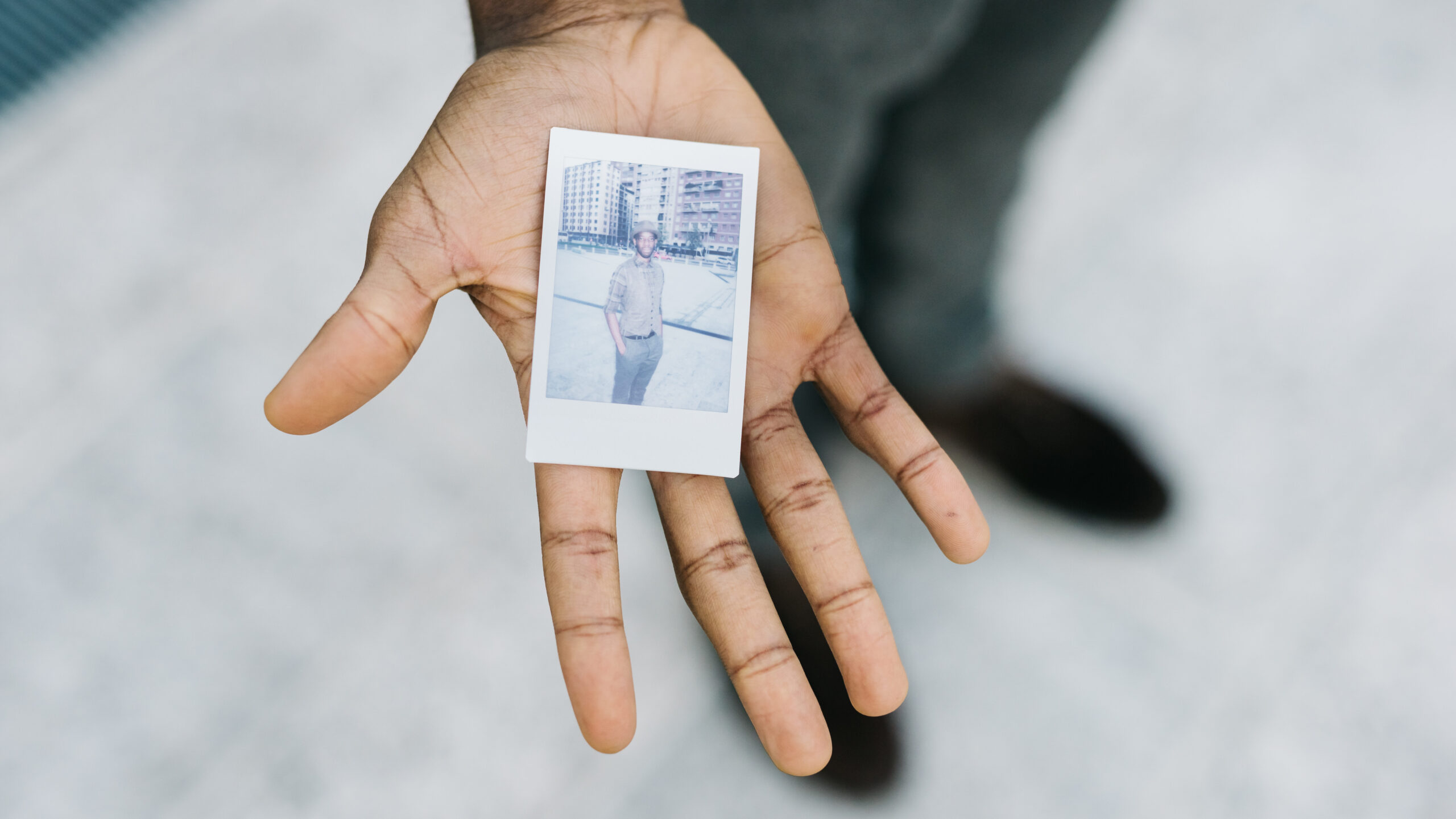 Imagem de uma mão segurando um retrato desbotado em polaroide de um homem negro com roupa social esportiva e chapéu.