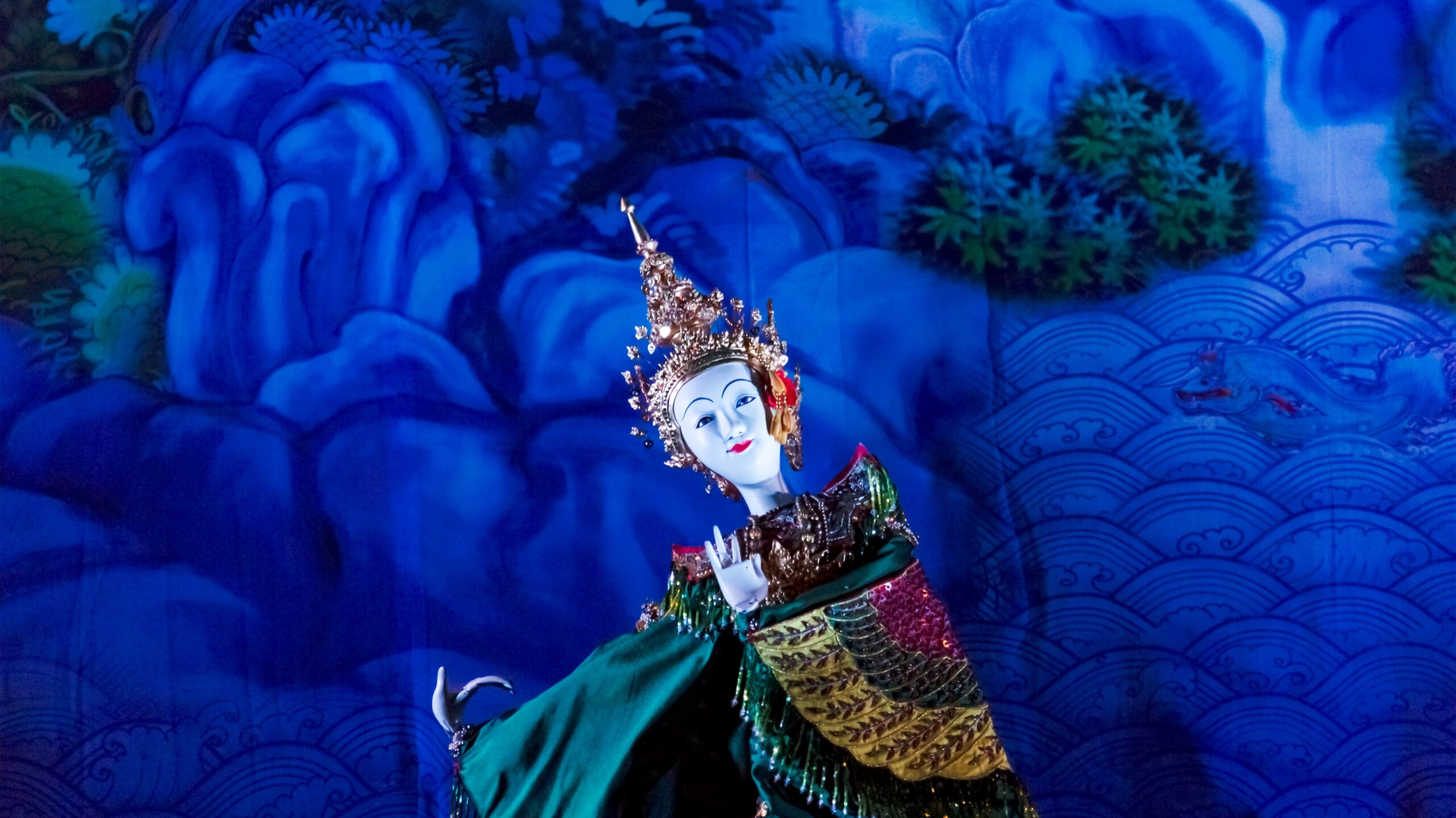 Fantoche tradicional tailandês vestido com trajes elaborados e coloridos, incluindo um adorno dourado na cabeça e brincos vermelhos. Ele está posicionado diante de um fundo azul vibrante, ricamente detalhado com padrões que lembram ondas e folhagens, criando uma cena mística e abstrata.